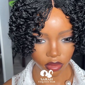 Puede incluir: Una peluca negra con un estilo rizado y trenzado. La peluca está en una cabeza de maniquí con una blusa de encaje blanco y un collar de perlas. El texto "SARAH EXQUISITE HAIR" es visible debajo del cuello del maniquí.