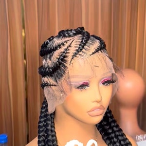 Puede incluir: Una peluca negra con el pelo trenzado en un patrón de trenza de corona. La peluca está en una cabeza de maniquí con un frente de encaje. La peluca está diseñada para permitir una línea de cabello natural. La peluca está etiquetada como "SARAH EXQUISITE HAIR".