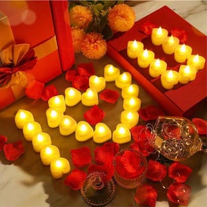 Könnte beinhalten: Eine Herzform aus kleinen, gelben, batteriebetriebenen LED-Teelichtern auf einem Tisch mit roten Rosenblättern. Die Lichter sind in Herzform angeordnet und von roten Rosenblättern umgeben. Im Hintergrund befinden sich eine rote Geschenkbox und eine Glasvase mit einer Lichterkette.