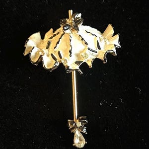 May include: A white and black enamel umbrella brooch with a rhinestone accent. The umbrella has a black bow at the bottom and is decorated with rhinestones.