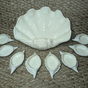 Könnte beinhalten: Eine große, cremeweiße Servierschale aus Keramik in Form einer Muschel, begleitet von acht kleineren, passenden muschelförmigen Schälchen. Jedes Stück hat eine glatte, glänzende Oberfläche und realistische, geformte Texturen.