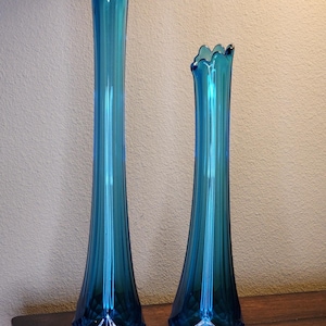 May include: Two tall, turquoise glass vases with ruffled rims. The vases have a slender, ribbed design and diamond-shaped bases. One vase is taller than the other. The vases are displayed on a dark wooden surface.