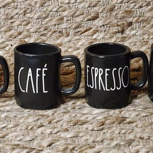 May include: Four black ceramic coffee mugs with white text. Two mugs say "CAFÉ" and two say "ESPRESSO". The mugs have a simple, rounded shape with a handle. The mugs are arranged on a woven, textured surface.