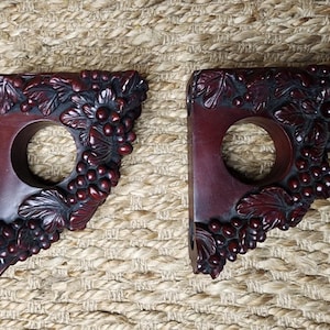 May include: Two dark red wooden decorative brackets with a carved grape and leaf design. Each bracket has a circular opening. The brackets are displayed on a textured, neutral-toned surface.
