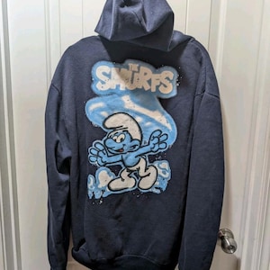 Könnte beinhalten: Marineblauer Kapuzenpullover mit einem Schlumpf-Motiv und dem Schriftzug "The Smurfs" in Weiß und Hellblau. Der Pullover hat eine Kapuze mit Kordelzug und eine Kängurutasche.