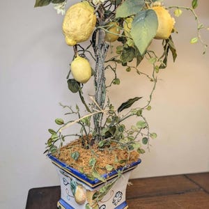 May include: A decorative artificial lemon tree with yellow fruit, green leaves, and small white flowers. The tree is planted in a square white ceramic planter featuring blue trim and painted lemon motifs on the exterior sides.
