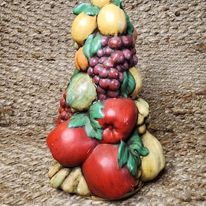 Könnte beinhalten: Eine Keramik-Fruchtskulptur mit einer Vielzahl farbenfroher Früchte. Die Skulptur umfasst rote Äpfel, lila Trauben, gelbe Zitronen und grüne Birnen. Die Früchte sind in einem gestapelten, dekorativen Design mit grünen Blättern angeordnet.
