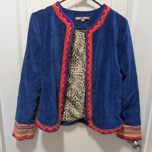 May include: A blue velvet jacket with a leopard print lining. The jacket features colorful embroidered trim in shades of orange, pink, and gold along the edges and cuffs. The jacket is hanging on a black hanger against a white door.