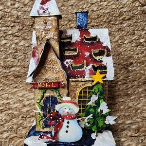 Puede incluir: Un portacalzas navideño decorativo con forma de casa festiva. La casa presenta un muñeco de nieve, un árbol de Navidad y la palabra "NOEL" en una pancarta. El tejado es rojo con detalles blancos y la base es azul.