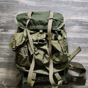 Könnte beinhalten: Ein olivgrüner Rucksack im Militärstil mit einer großen oberen Klappe, mehreren Außentaschen mit Druckknöpfen und langen, verstellbaren Gurtbändern. Der untere Rahmenbereich ist sichtbar, auf dem das Wort LONG aufgedruckt ist.