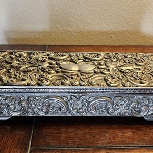 Puede incluir: Una caja rectangular de metal plateado con un intrincado patrón en relieve de flores y volutas en la tapa y los laterales. La caja se apoya sobre cuatro patas pequeñas y presenta un acabado metálico texturizado con detalles oscuros.