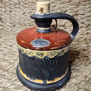 Op de afbeelding: Een handgemaakte keramische karaf met een kurken stopper. De karaf heeft een rode bovenkant, een zwarte handgreep en een zwarte basis met een getextureerd, geribbeld ontwerp. De basis is versierd met gele en groene accenten.