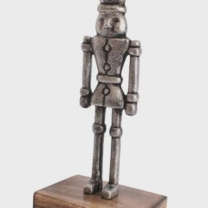 Puede incluir: Una figura de cascanueces de metal plateado se encuentra sobre una base de madera con un gancho. El cascanueces tiene un diseño detallado, que incluye un sombrero, una chaqueta y piernas. La base de madera es rectangular.