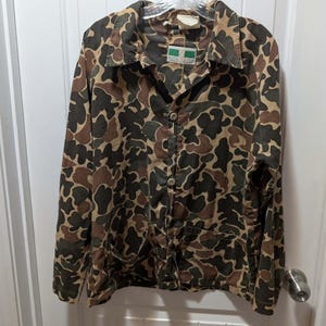May include: A vintage camouflage jacket with a button-up front and two front pockets. The jacket features a brown, green, and tan pattern. The jacket is hanging on a white door.