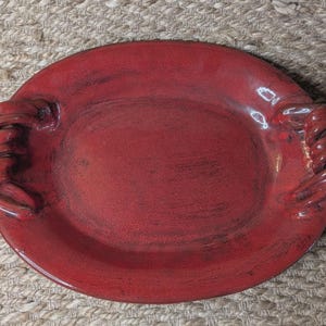 May include: An oval-shaped ceramic serving platter finished in a deep, glossy red glaze with a subtle mottled texture. The platter features two integrated handles on opposite sides, each designed with a distinct, thick, coiled rope-like appearance.