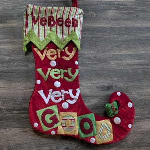 May include: A red Christmas stocking with the words "I've Been Very Very Very Good" in various colors. The stocking has a green and white striped top and a green zig-zag trim. White polka dots and a small green gift adorn the stocking.