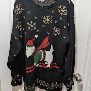 Könnte beinhalten: Schwarzer Strickpullover mit goldenen Schneeflocken und einem Cartoon-Hund mit Weihnachtsmütze und Schal. Der Pullover hat einen gewellten Saum und Manschetten mit grünen und goldenen Details, ideal für die Weihnachtszeit.