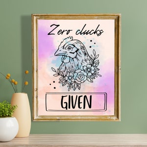May include: A framed print with a watercolor background in shades of pink, blue, and purple. A black and white illustration of a chicken's head with flowers around it. The text "Zero Clucks Given" is printed in black.