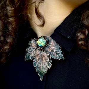 May include: A copper-toned metal brooch shaped like a maple leaf with a textured, veined surface. A square-cut labradorite gemstone with iridescent green and blue flashes is secured at the center of the leaf by a decorative copper wire wrap.