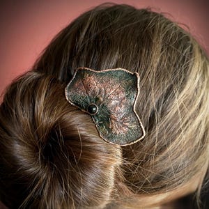 Op de afbeelding: Een haarclip in de vorm van een blad in kopertinten, met een donkergroene en bronzen afwerking. De clip heeft een kleine, ronde, zwarte steen in het midden. De clip is bevestigd aan een knot van bruin haar.