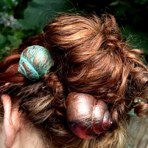 May include: Two snail shell hair accessories secured in auburn hair. One shell features a textured turquoise and copper patina finish, while the other displays a smooth, metallic copper and bronze gradient. Both shells are spiraled and rounded.