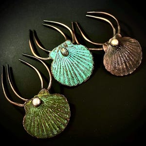 Puede incluir: Tres horquillas para el cabello con diseños de conchas de vieira de metal. Una concha es turquesa con una piedra central, una es bronce oscuro con una perla blanca y una es verde oliva con una perla blanca. Cada concha tiene dos púas curvas.