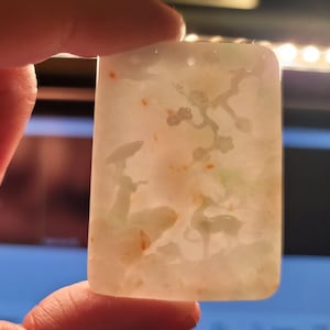 May include: A white jade pendant with a carved design of a person holding an umbrella and a deer standing in a field. The jade has a light green hue and is translucent.