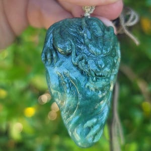 May include: A green jade pendant carved in the shape of a dragon. The dragon is curled up with its head facing forward. The pendant is hanging from a cord.