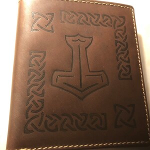 Puede incluir: Cartera de cuero marrón con un borde de nudo celta y un diseño de martillo Mjolnir en el centro.