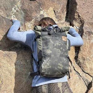 以下が含まれることがあります： グレーのシャツと迷彩柄のレギンスを着用した人が岩壁を登っています。黒と迷彩柄のバックパックを背負っており、バックパックには「Mystery Ranch」と書かれた茶色の革のパッチが付いています。