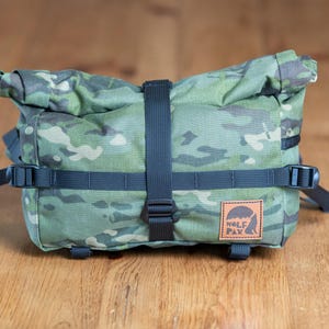 May include: A green and brown camouflage bike handlebar bag with black straps and a brown leather patch with the text "Wolf Pax" in black.