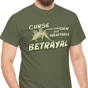 Può includere: T-shirt verde oliva con la scritta "Curse Your Sudden But Inevitable Betrayal" e un dinosauro dei cartoni animati. Il design è in stile vintage con un aspetto invecchiato. La maglietta è girocollo e a maniche corte.