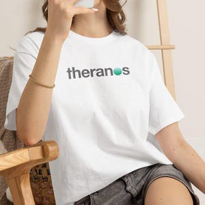 Op de afbeelding: Een wit T-shirt met korte mouwen waarop het woord theranos in donkergrijze kleine letters is gedrukt, waarbij een groenblauwe cirkel de letter o vervangt. Het shirt wordt gecombineerd met een donkergrijze spijkerbroek voor een casual outfit.