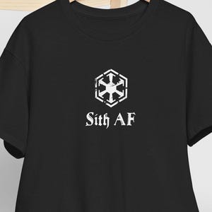 May include: A black short-sleeved t-shirt featuring a white distressed hexagonal emblem with a central star-like symbol and the text "Sith AF" printed in a gothic-style font. The shirt is displayed on a wooden hanger alongside a plain white baseball cap.