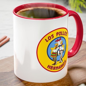Peut inclure: Tasse en céramique blanche avec intérieur et anse rouges, arborant le logo "Los Pollos Hermanos". La tasse contient du café chaud et est posée sur une planche à découper en bois. Bâtons de cannelle et pomme de pin visibles.