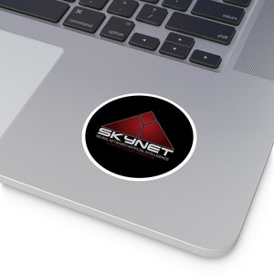 May include: A black and red circle sticker with the text "SKYNET" in white letters. The text "NEURAL NET-BASED ARTIFICIAL INTELLIGENCE" is written in smaller white letters below the logo.
