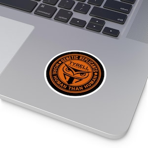 May include: A round sticker with the text "MORE HUMAN THAN HUMAN" and "GENETIC REPLICANTS TYRELL CORPORATION". The sticker has an orange and black color scheme, featuring an owl design in the center.