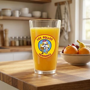 May include: A pint glass filled with orange juice, displaying a circular logo with the words "Los Pollos Hermanos" and two cartoon chickens. The glass is on a wooden surface, with a bowl of fruit in the background.
