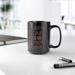 May include: A black coffee mug with the word "COFFEE" in multiple languages printed in brown. The mug is on a white surface with a black pen and a notebook in the background.