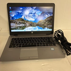 May include: A silver HP EliteBook laptop with a black keyboard, a trackpad, and a screen displaying a mountain landscape. A black power adapter with a coiled cord is positioned to the right of the laptop. The device features an Intel Core processor sticker.