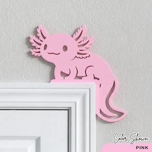 Peut inclure: D&eacute;coration rose en forme d'axolotl, plac&eacute;e sur le coin d'un cadre de porte blanc. L'axolotl a des branchies d&eacute;taill&eacute;es et un visage souriant. Le texte "Color Shown: PINK" est dans le coin inf&eacute;rieur droit.
