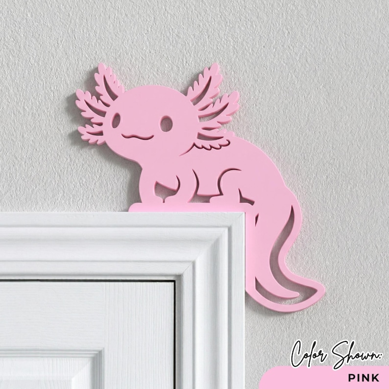Peut inclure: D&eacute;coration rose en forme d'axolotl, plac&eacute;e sur le coin d'un cadre de porte blanc. L'axolotl a des branchies d&eacute;taill&eacute;es et un visage souriant. Le texte "Color Shown: PINK" est dans le coin inf&eacute;rieur droit.