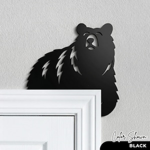 May include: A matte black metal wall art piece shaped like a bear, designed to be positioned in the corner of a door frame. The silhouette features cut-out details along the fur and face, creating a distinct outline of a bear looking upward.