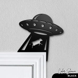 May include: Black metal wall art depicting a UFO beaming up a cow. The UFO has a classic saucer shape with portholes. The cow is shown mid-air, being lifted by the UFO's tractor beam. The text "Color Shown: BLACK" is at the bottom.