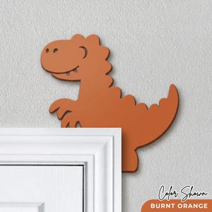 May include: Burnt orange dinosaur wall decor. The dinosaur has a friendly expression with a curved smile and two small teeth. The color shown is burnt orange. The dinosaur is mounted on a white wall.
