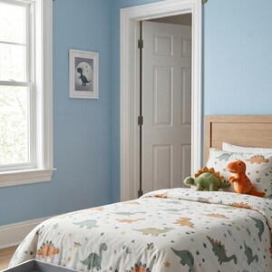 Puede incluir: Un dormitorio infantil con una pared azul claro, una cama con una funda n&oacute;rdica con tem&aacute;tica de dinosaurios y una cabecera de madera. Recortes de dinosaurios decoran la pared. Una imagen enmarcada de un dinosaurio est&aacute; en la pared. El texto "LUCKYBORDERS.ETSY.COM" est&aacute; en la parte inferior.