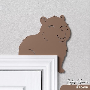May include: A brown capybara wall decoration designed to sit on the top corner of a door frame. The flat, cutout silhouette features a smiling face with closed eyes and detailed fur markings, rendered in a solid matte brown finish throughout.