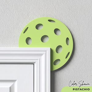 May include: A pistachio green, semi-circular door stop with circular cutouts, positioned above a white door frame. The text "Color Shown: Pistachio" is displayed in the lower right corner. The door stop is designed to prevent a door from slamming shut.