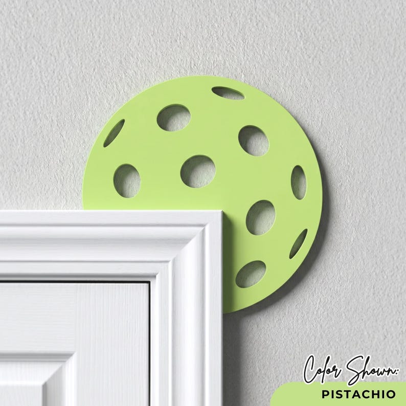 May include: A pistachio green, semi-circular door stop with circular cutouts, positioned above a white door frame. The text "Color Shown: Pistachio" is displayed in the lower right corner. The door stop is designed to prevent a door from slamming shut.