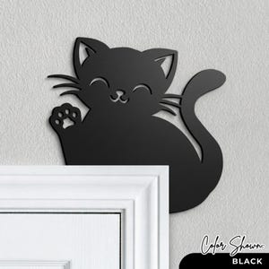 May include: A black cat-shaped wall decoration with a waving paw and a smiling face. The cat is cut from a solid material and mounted on a light gray wall above a white door frame. The text "Color Shown: BLACK" is at the bottom.